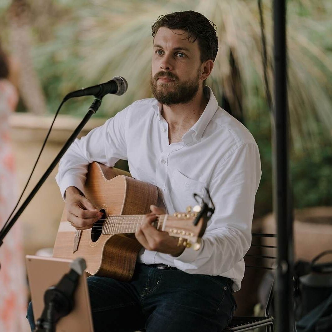 A bearded musician in a white shirt playing an acoustic guitar while singing into a microphone outdoors.