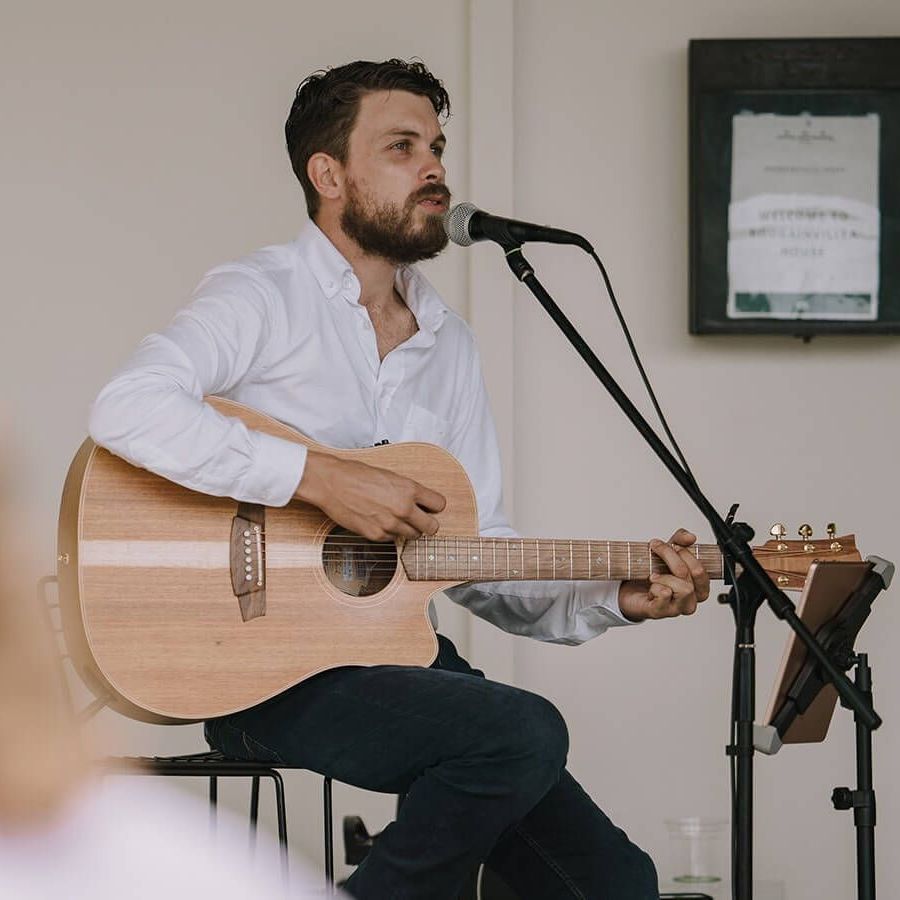 A person with a beard plays an acoustic guitar and sings into a microphone while seated indoors.