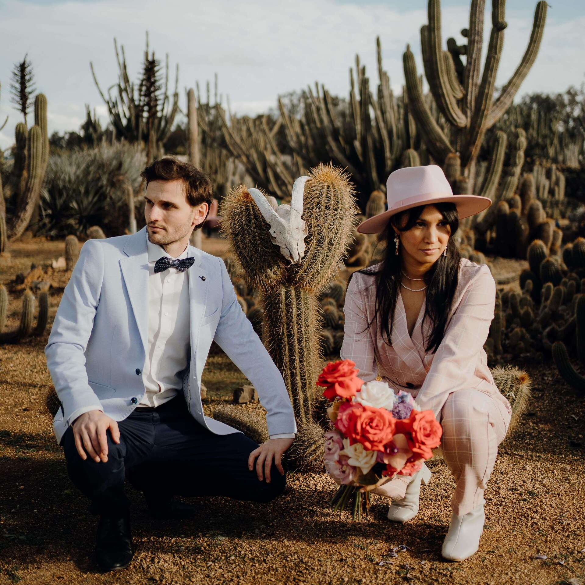 Couple in desert setting, man in light blue blazer, woman in pink suit holding bouquet, cacti in background.