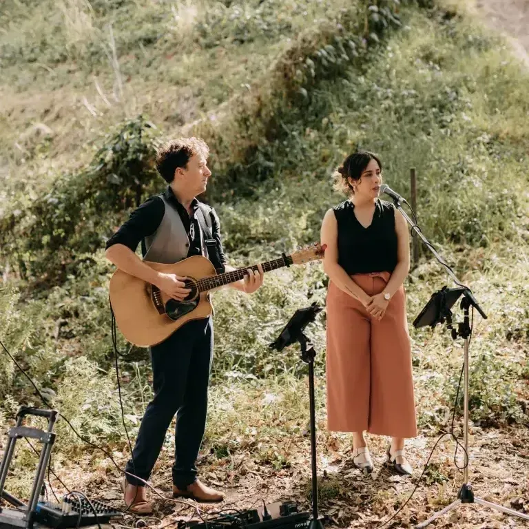 A duo performs outdoors: guitarist in vest and a singer in coral pants and black top.