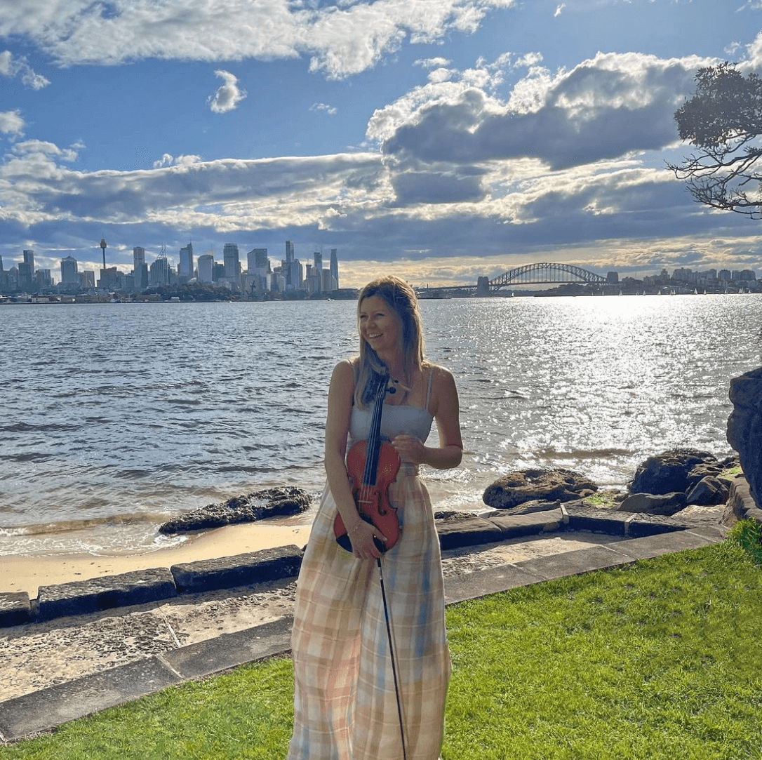 Woman holding a violin smiles on a grassy bank by water, cityscape in background. Blue sky, bright sunlight.