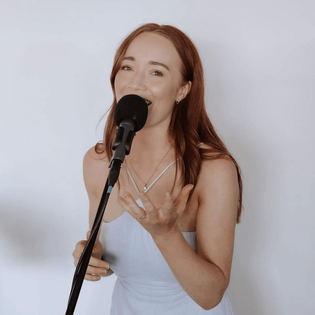 Woman with red hair singing into a microphone, wearing a light blue dress, white wall background.