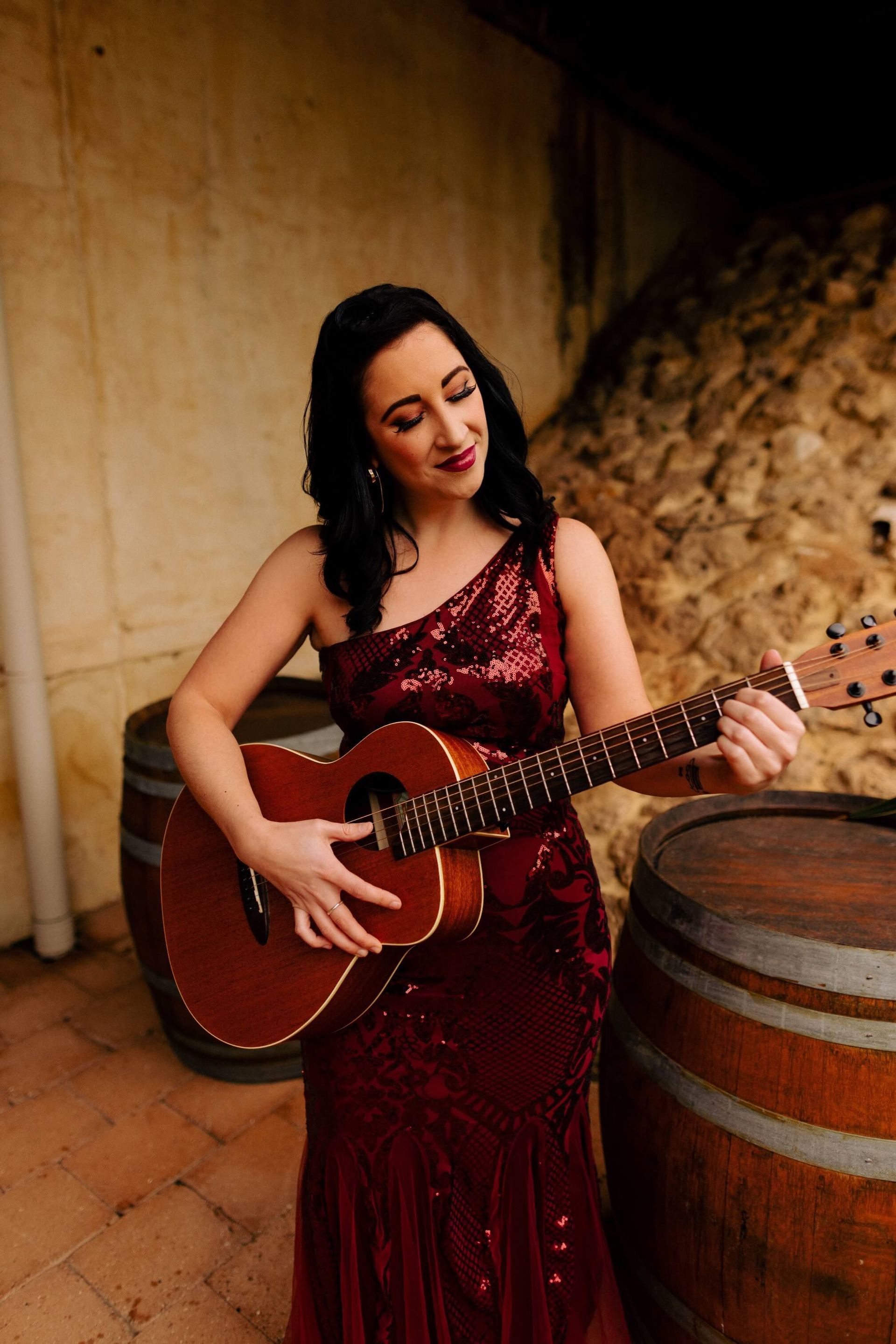 Woman in red sequined dress playing acoustic guitar near barrels.