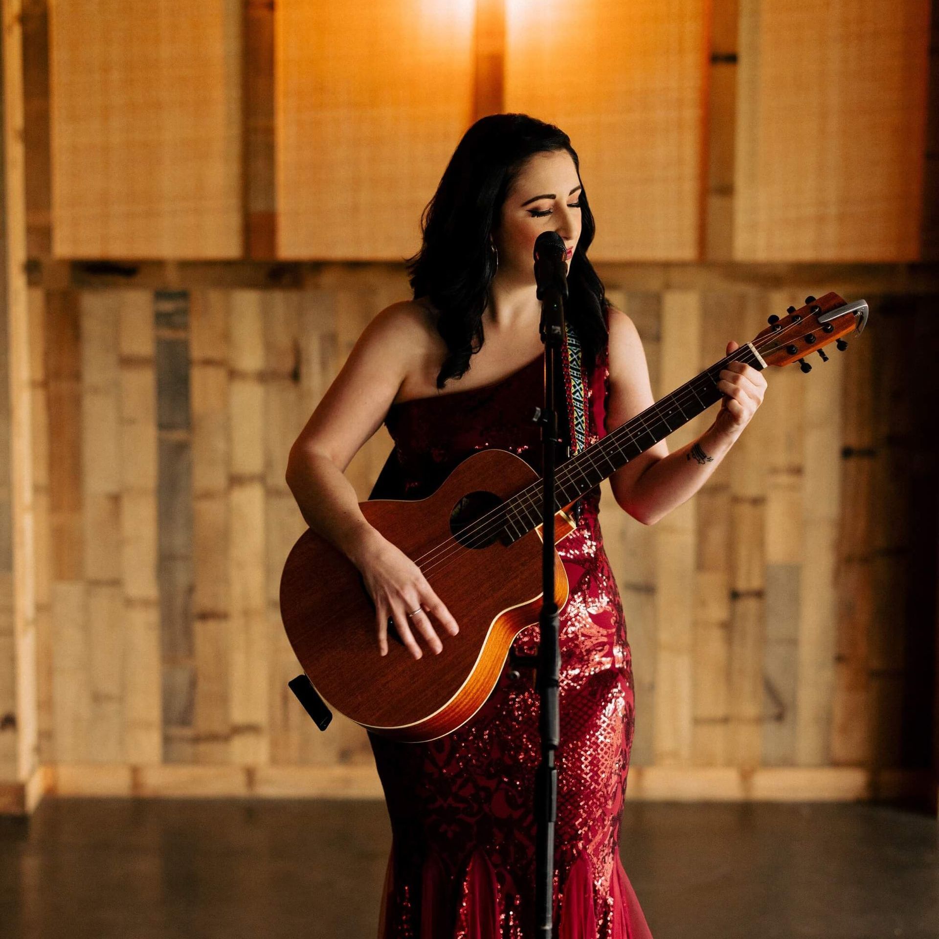 Woman in red dress plays acoustic guitar and sings into a microphone on stage.