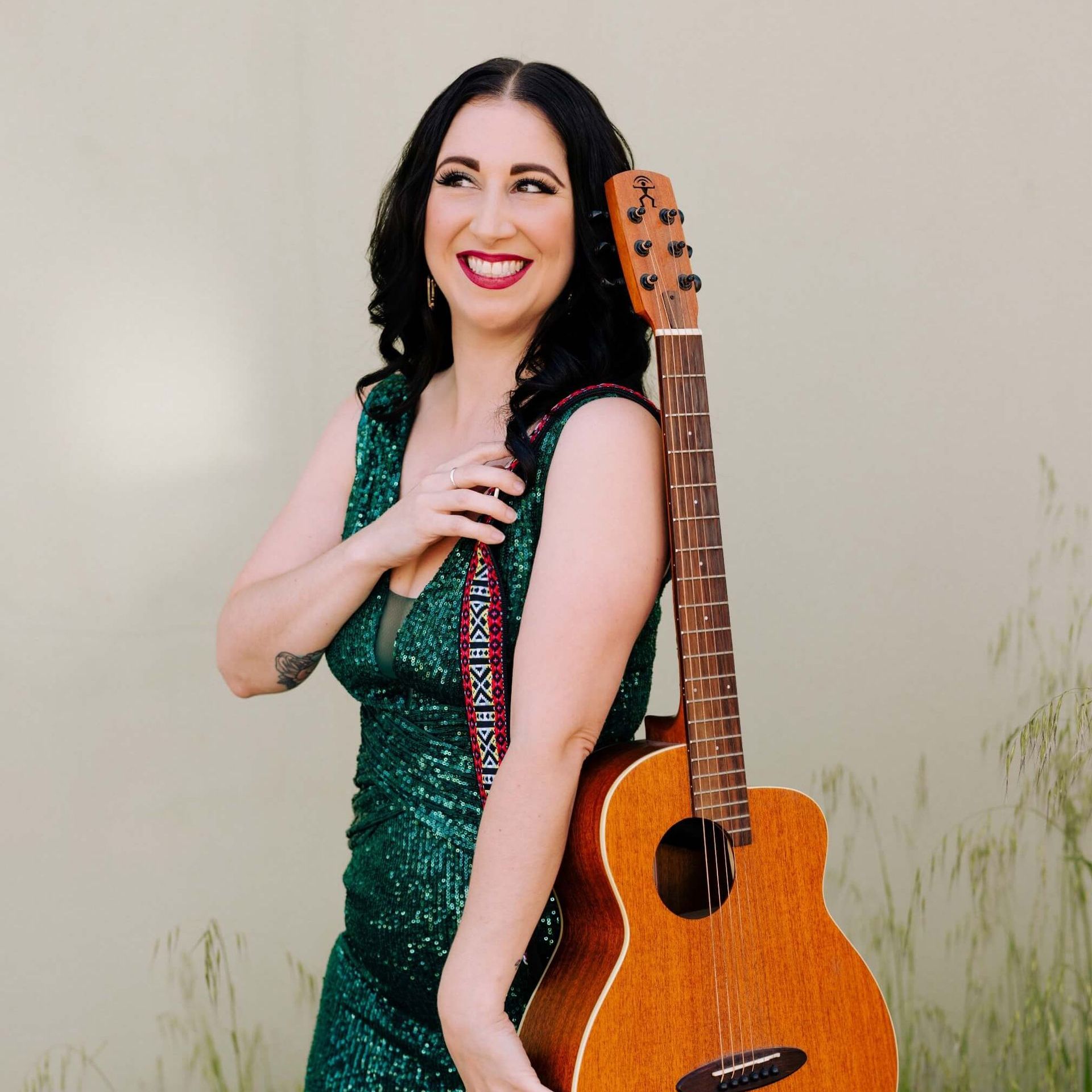 Woman in green sequined dress holds a brown acoustic guitar; smiles, leans against wall.