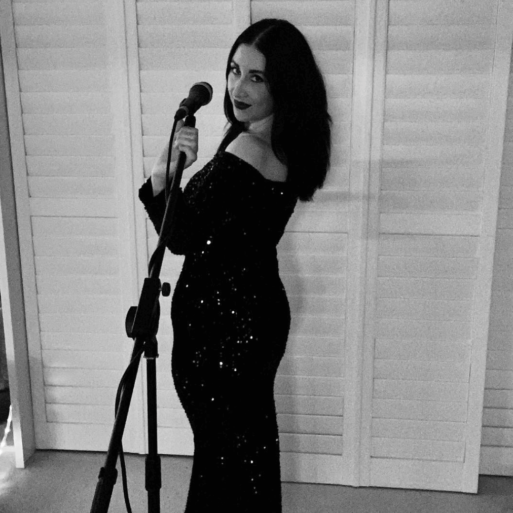 Woman in black sequined dress singing into a microphone. Standing in front of shuttered doors.