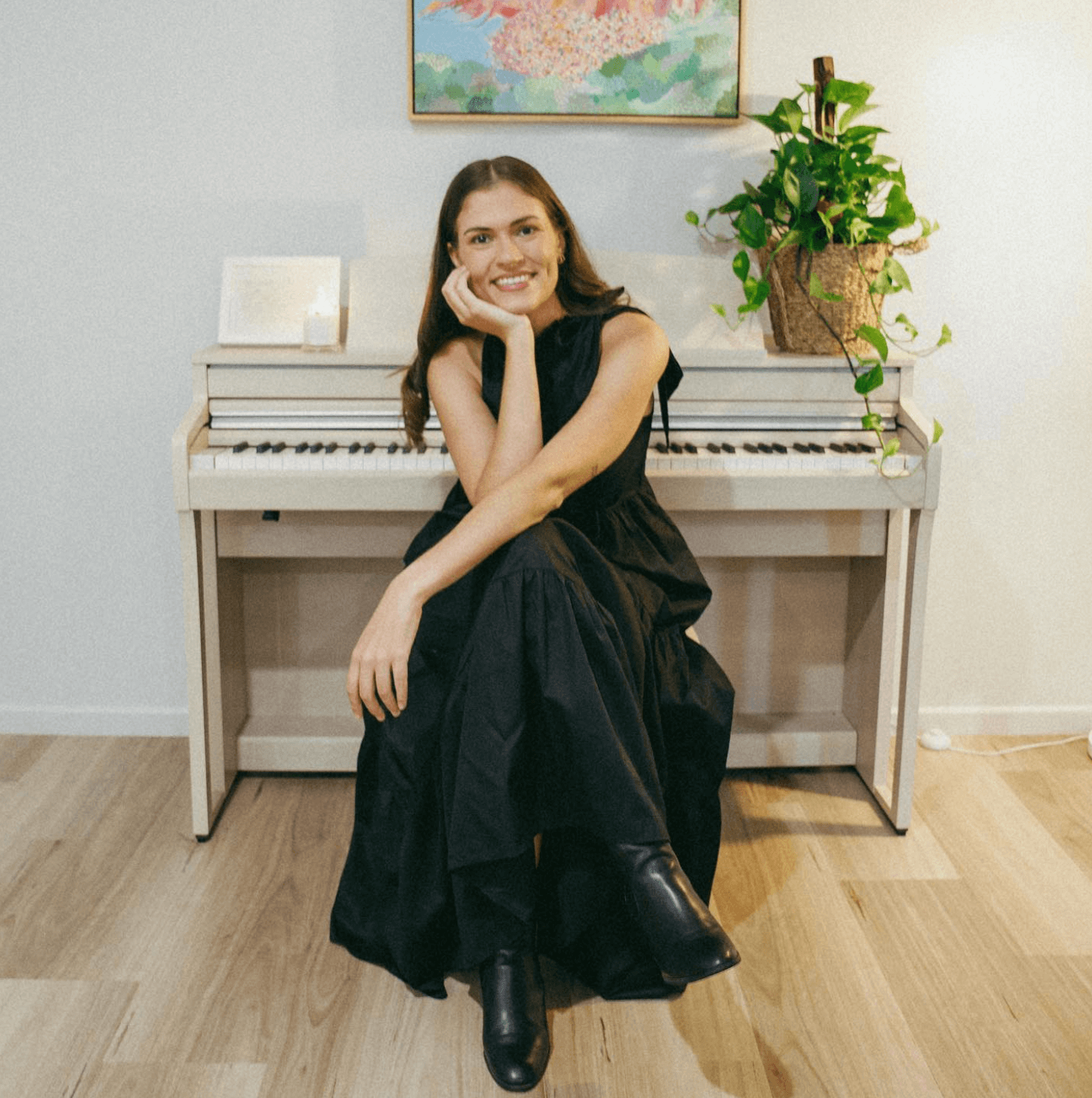 A smiling person in a black dress sits on a piano bench in front of an upright white piano with a potted plant on top.