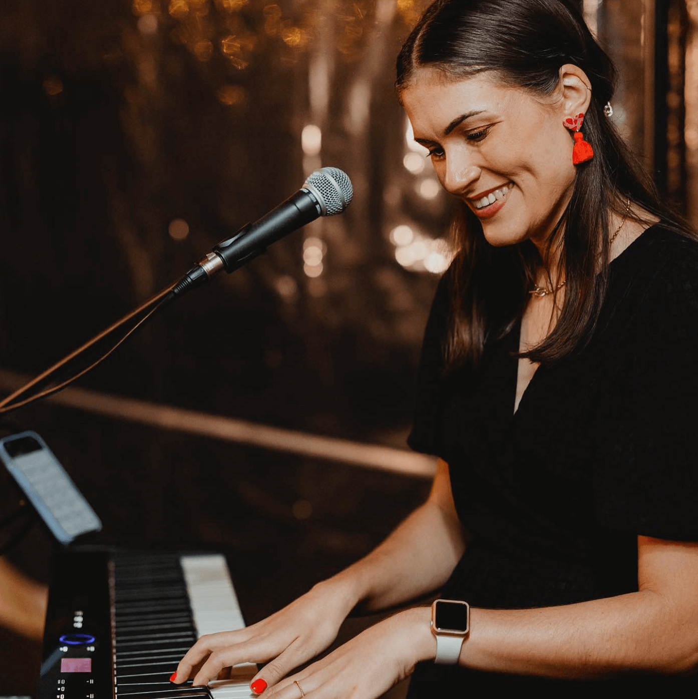 A person with dark hair and red earrings plays a keyboard while smiling, with a microphone positioned nearby.