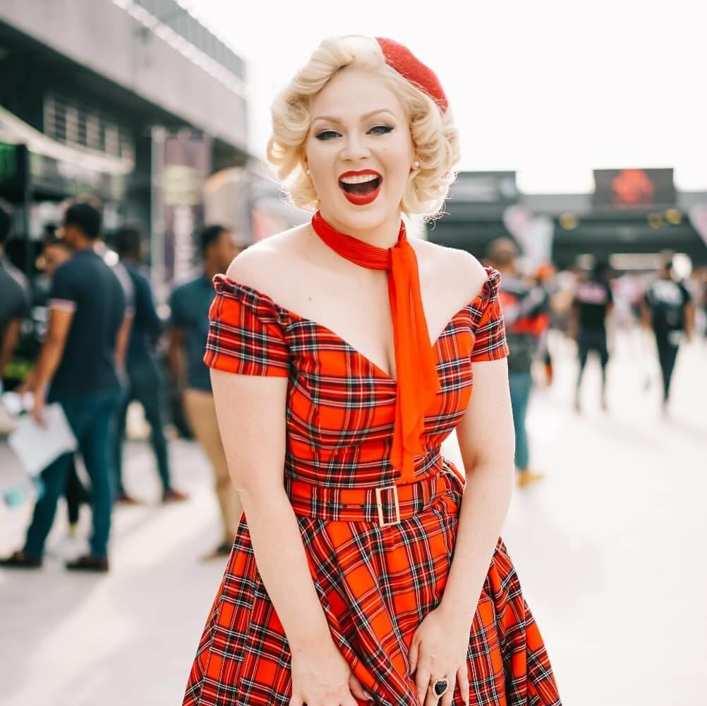 Woman in red plaid dress and accessories, smiling broadly outdoors.