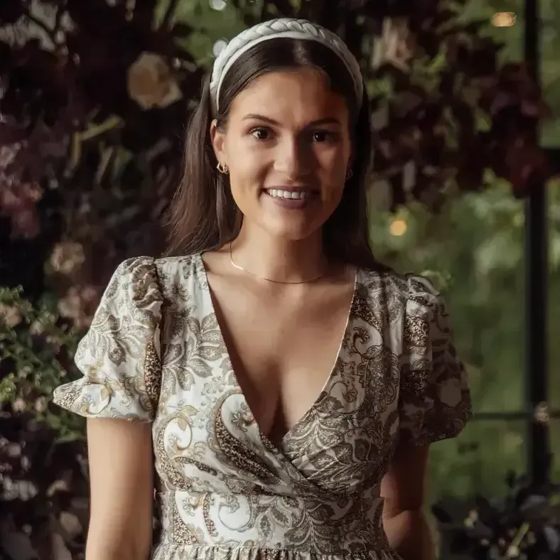 A person smiles wearing a white patterned headband and a brown and white paisley print dress against a floral background.