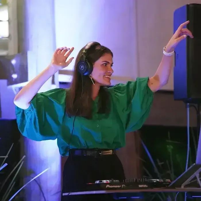 A person wearing a green blouse and headphones smiles while gesturing at a DJ console in a dimly lit event space.