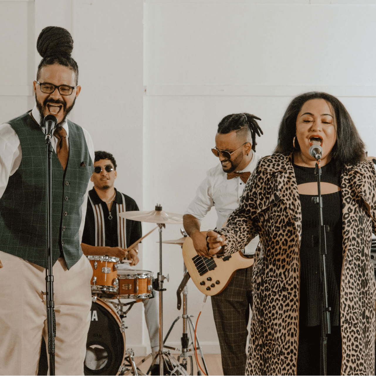 Band performing live; two vocalists, drummer, bassist; studio setting with white walls.