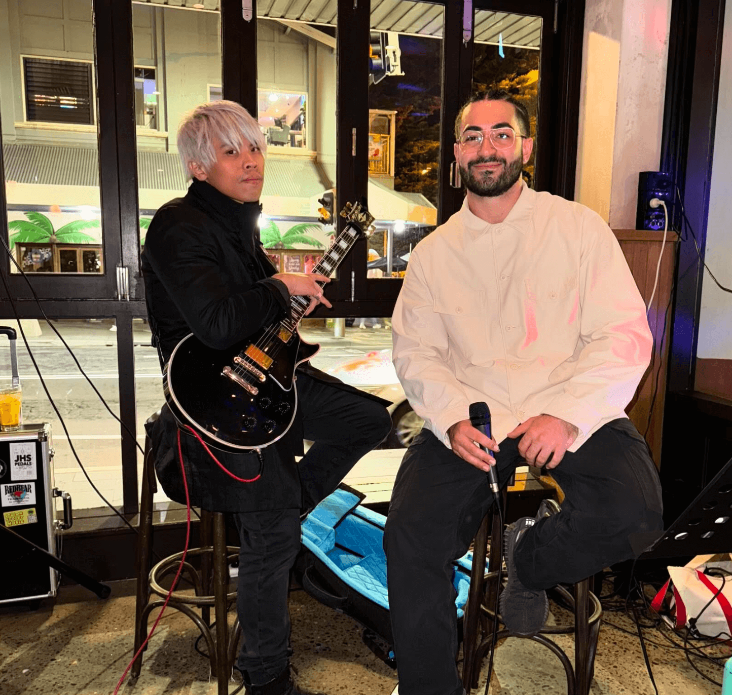 Two men are sitting on stools one is playing a guitar and the other is holding a microphone.