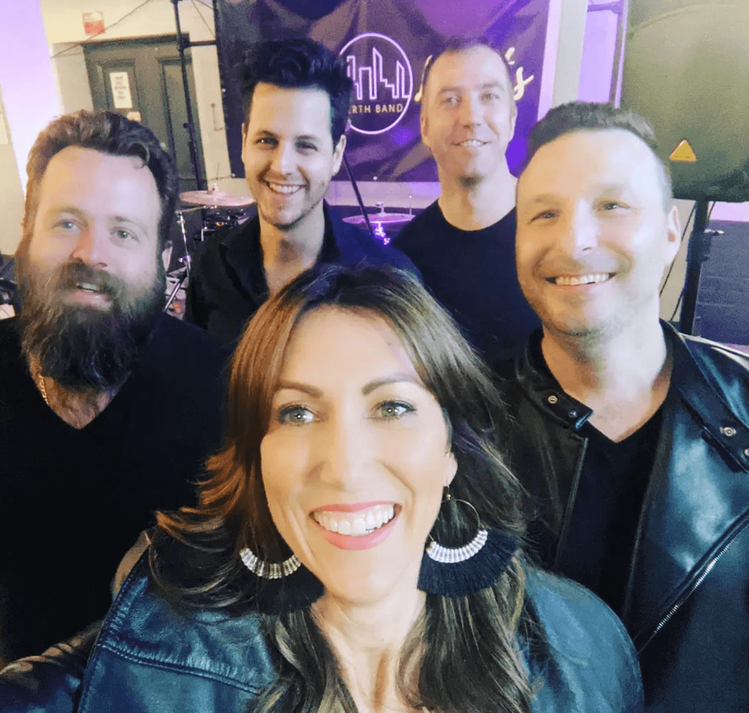 A group of five people smiling together in a selfie, likely a band posing in front of a logo backdrop.