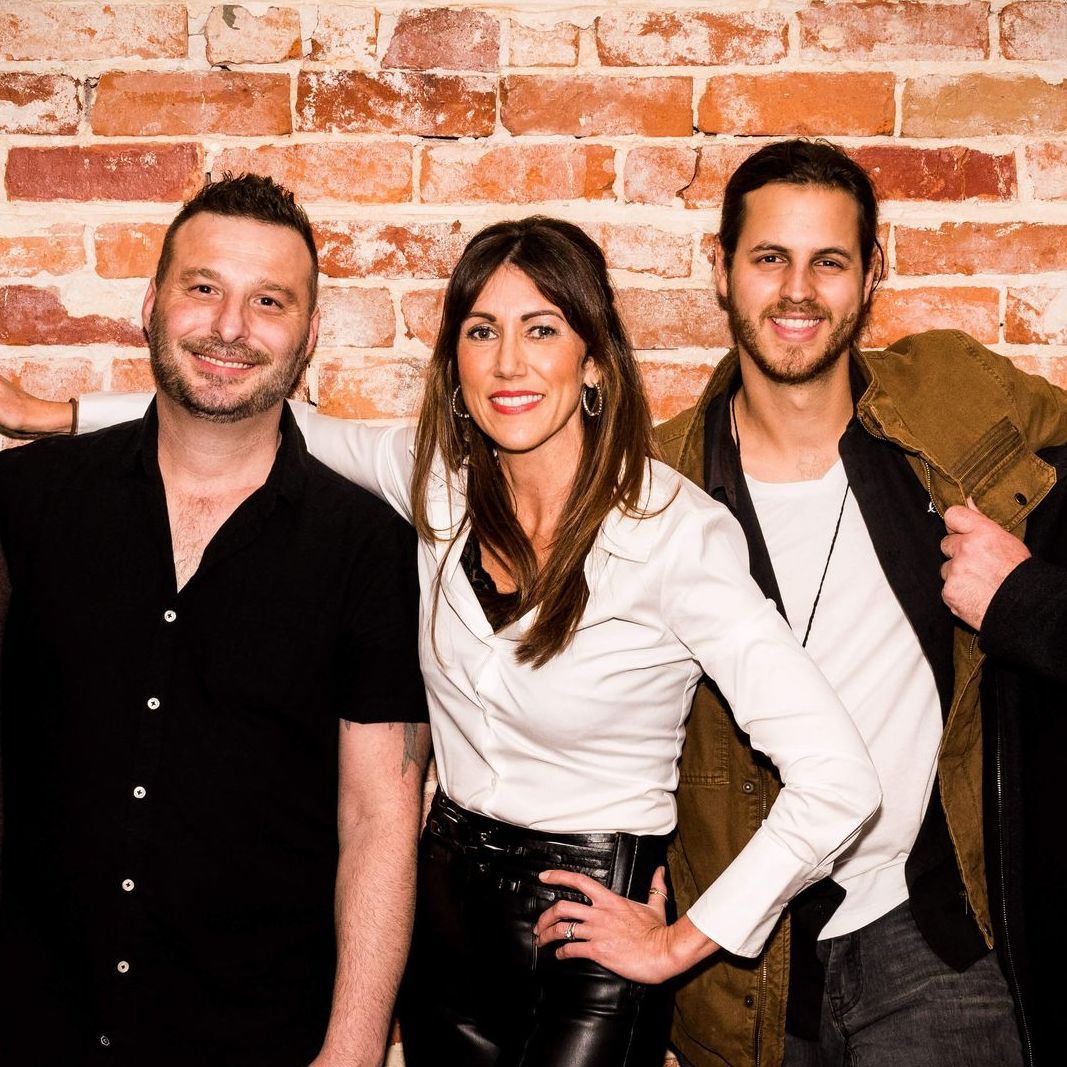 Three people stand side-by-side against a brick wall, posing for a photo with warm, friendly expressions.