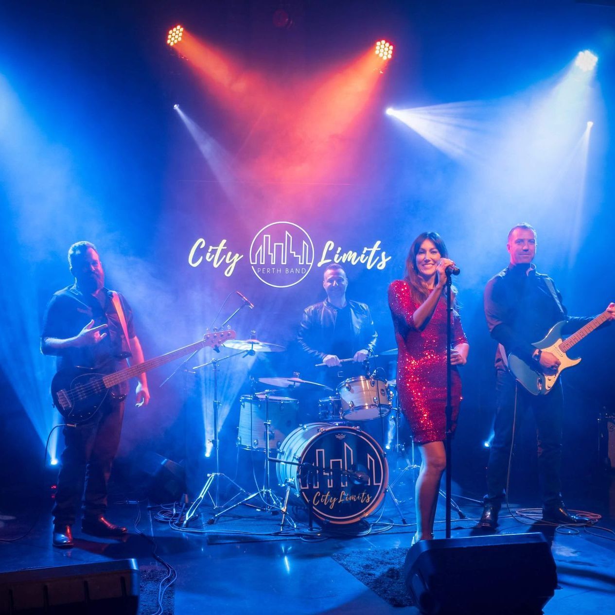 The band The Party Standard performs on a concert stage with red and blue spotlights, a singer in sequins, and instrumentals.