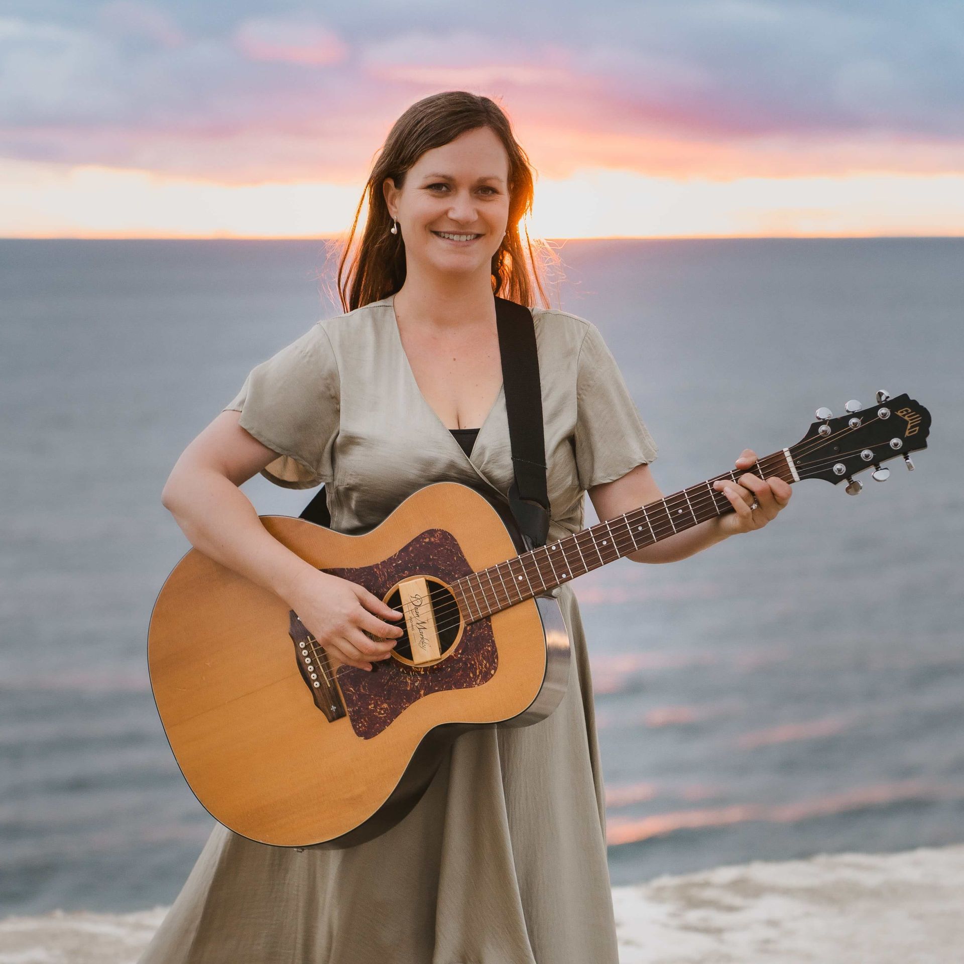 Woman playing acoustic guitar on a cliff overlooking the ocean at sunset.