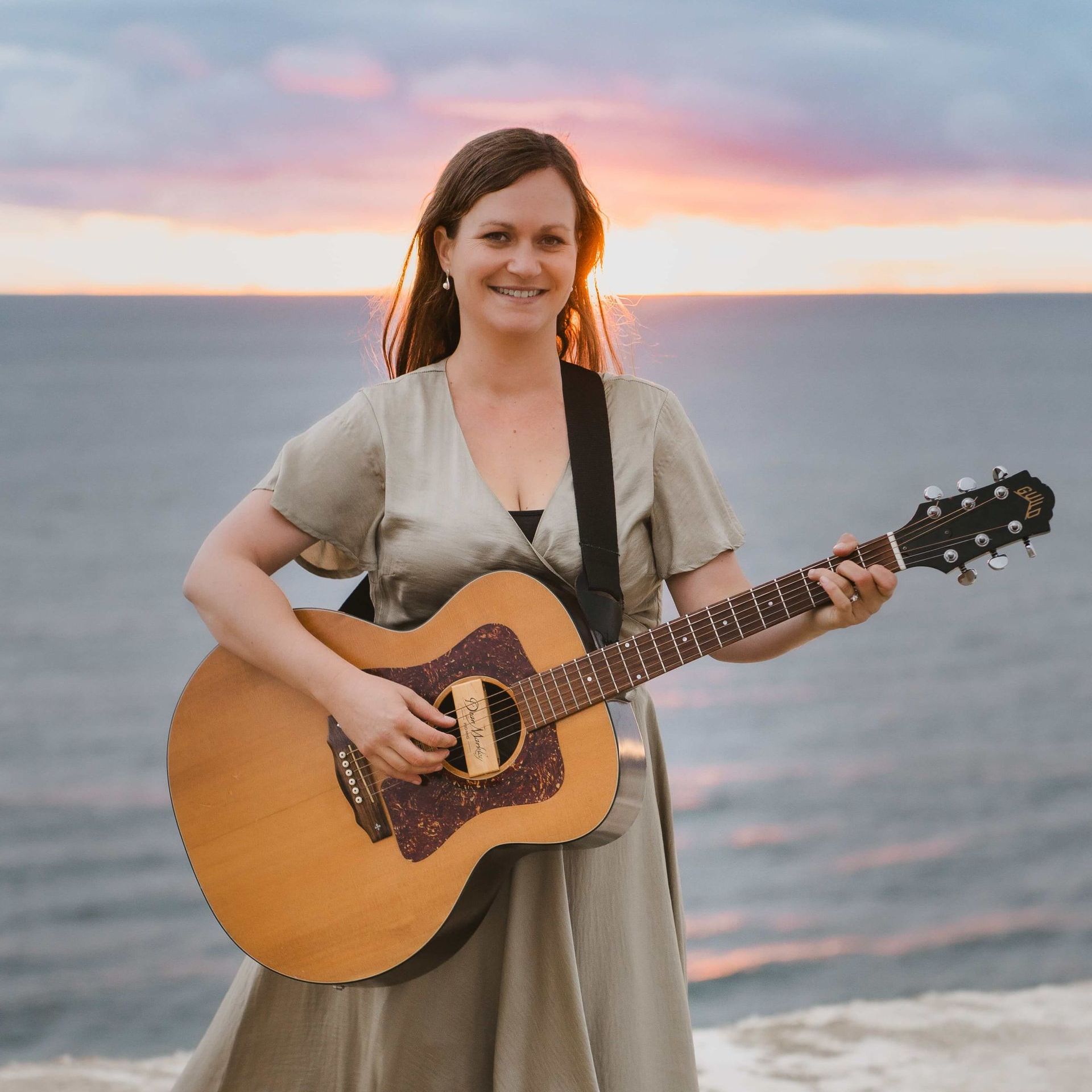 Woman with a guitar smiling at sunset near the ocean.