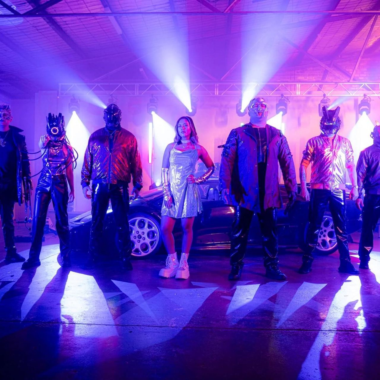 A woman in a silver dress stands in front of a black car, surrounded by figures in masks and shiny outfits under blue and purple lights.
