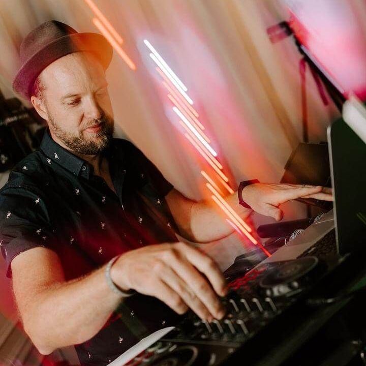 DJ mixing on turntables in a dim club, wearing a hat, with colorful blurred lights behind him