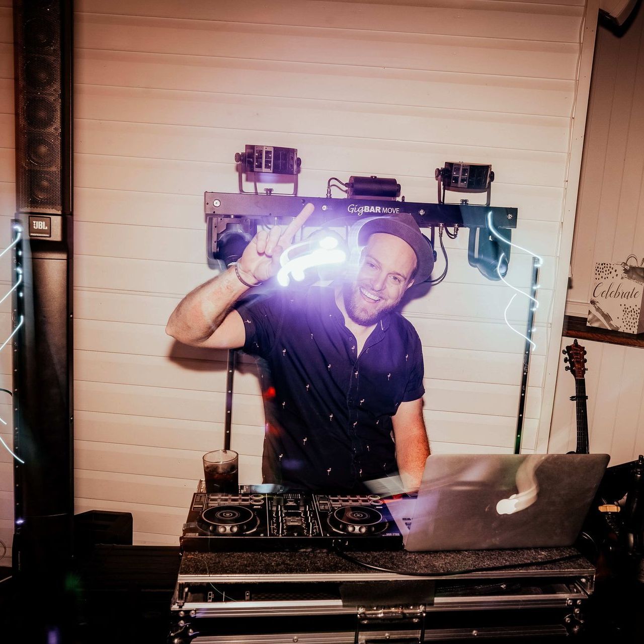 A smiling DJ in a dark shirt and hat gesturing with one hand behind a mixing console and laptop, illuminated by stage lights.