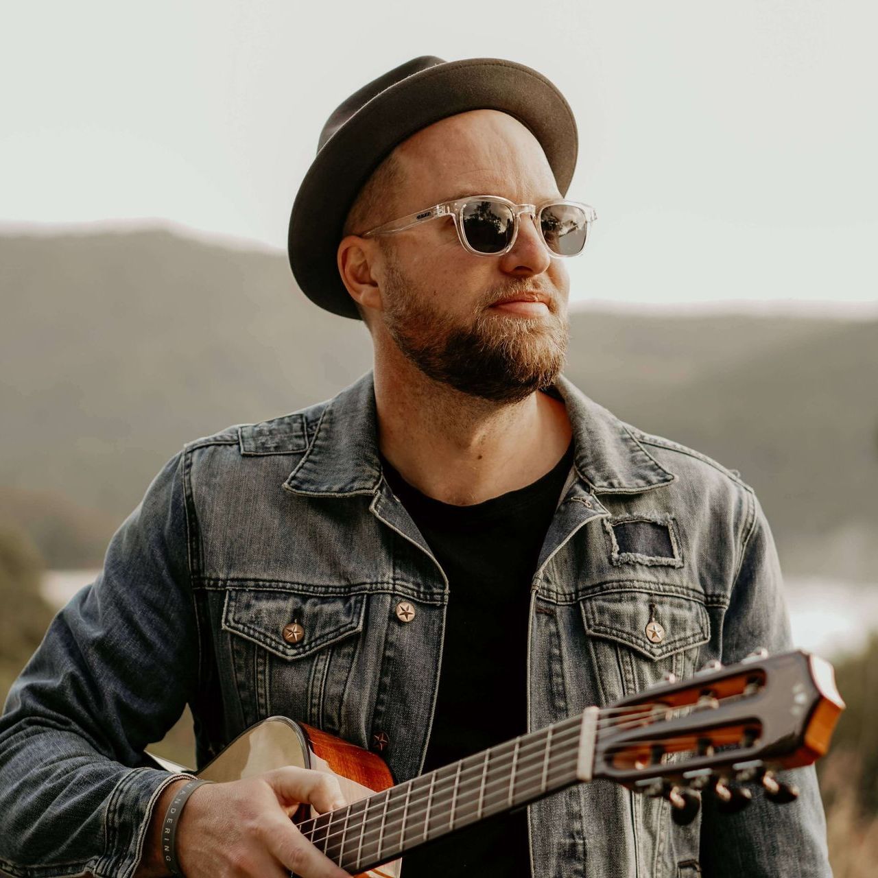 A person in a fedora, sunglasses, and denim jacket holds an acoustic guitar outdoors against a hilly, blurred background.