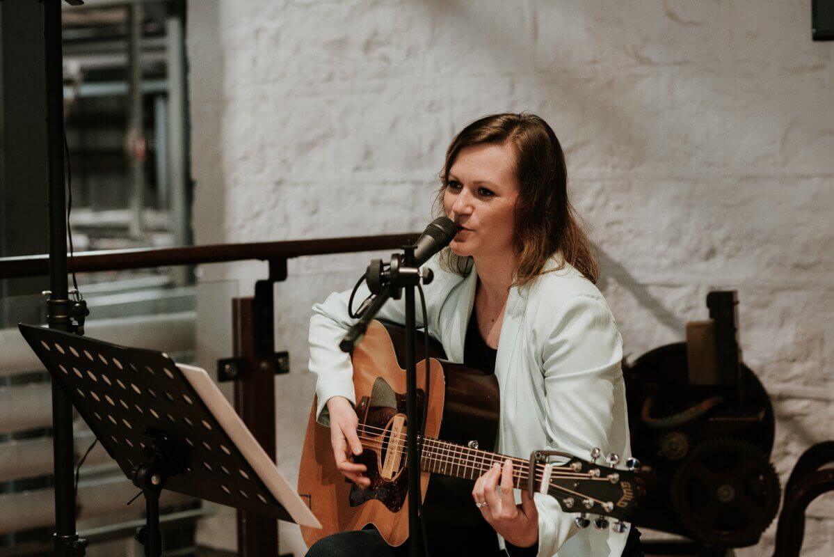 Woman playing acoustic guitar and singing into a microphone on stage, indoors, with a music stand.