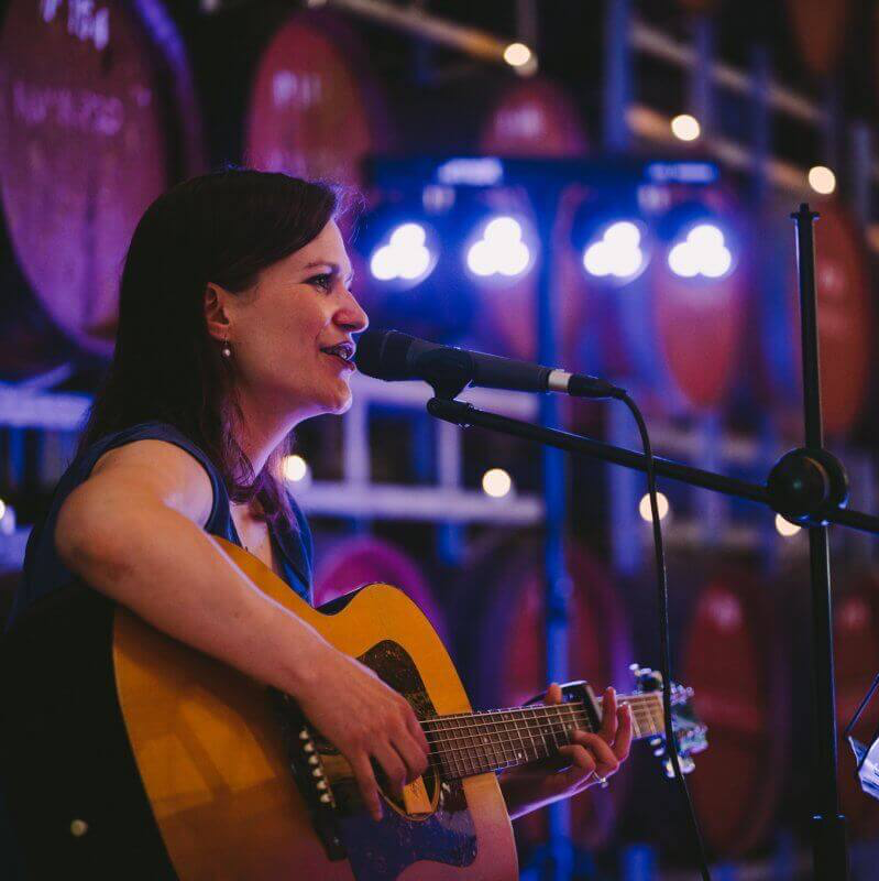 Woman playing guitar and singing into a microphone, lit by stage lights in front of wine barrels.