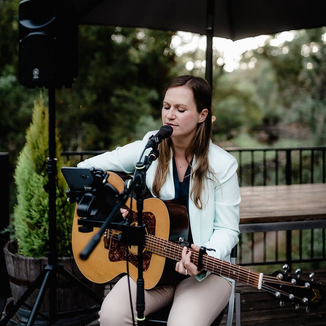 Woman playing acoustic guitar, singing into a microphone on an outdoor patio. She wears a light blue blazer.