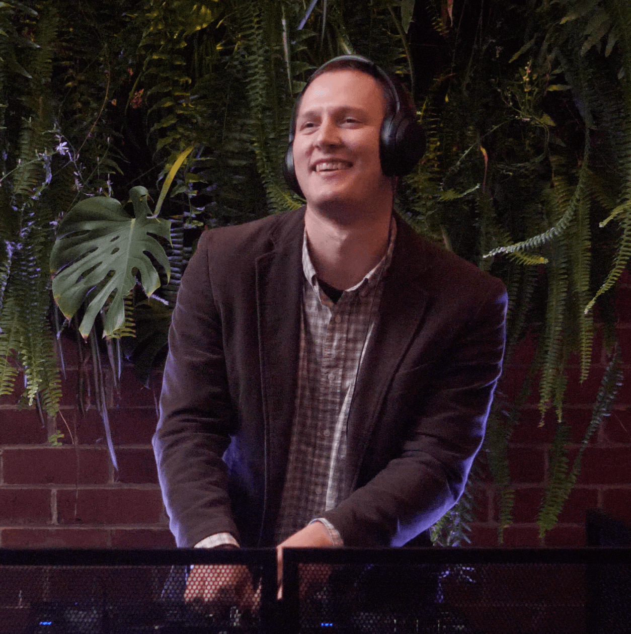 Man in a jacket and headphones smiles, standing near a brick wall with greenery in the background.