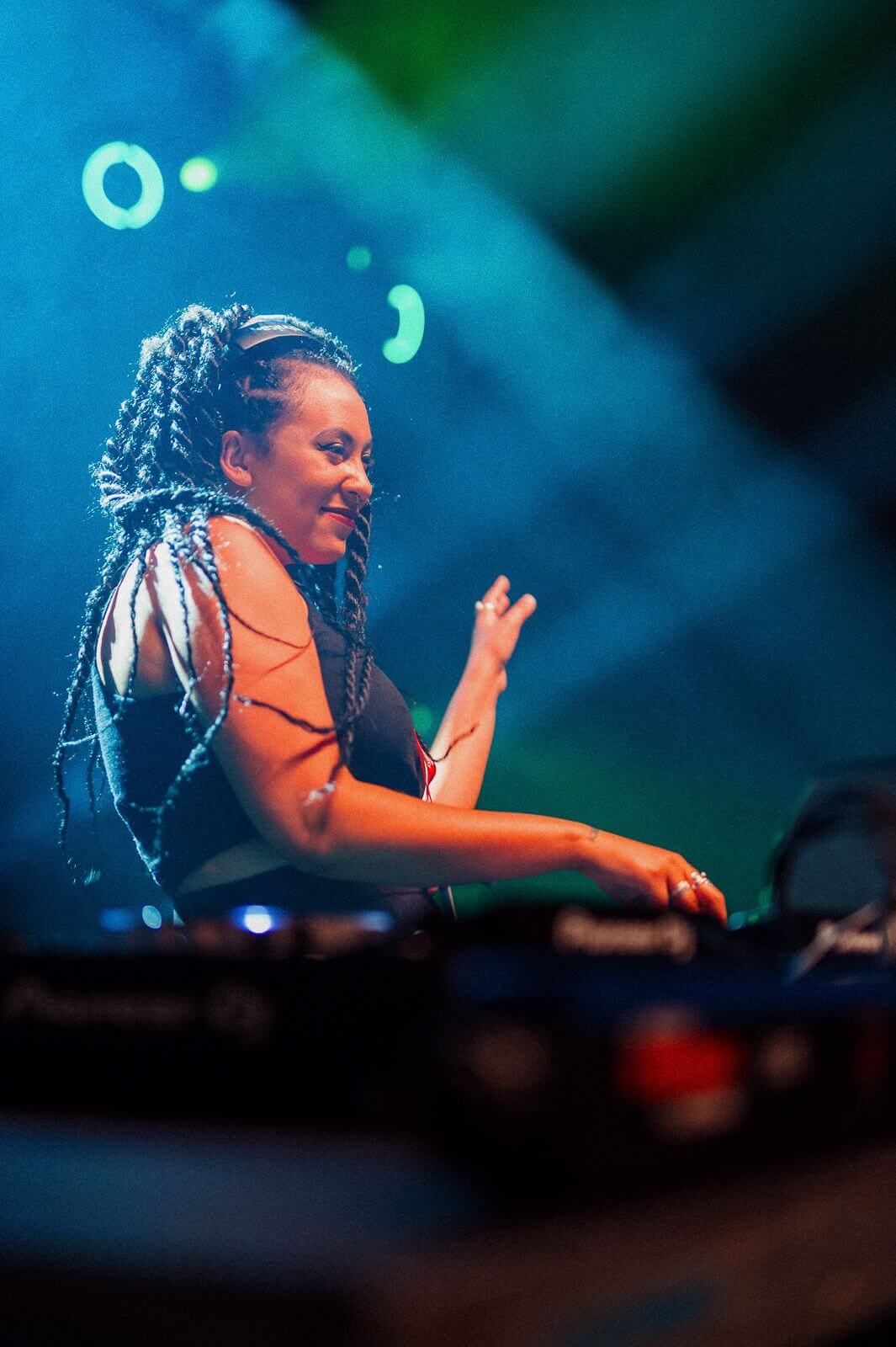 DJ performing on stage under blue lights, smiling at the mixer with braided hair and a sleeveless black top