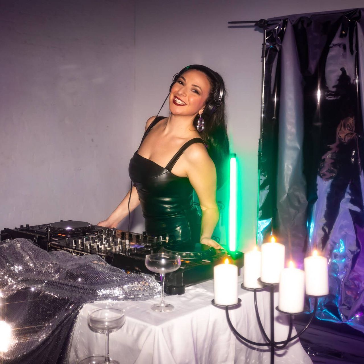 A DJ smiles while standing behind a mixing console at a table with candles and glassware in a dimly lit, festive setting.