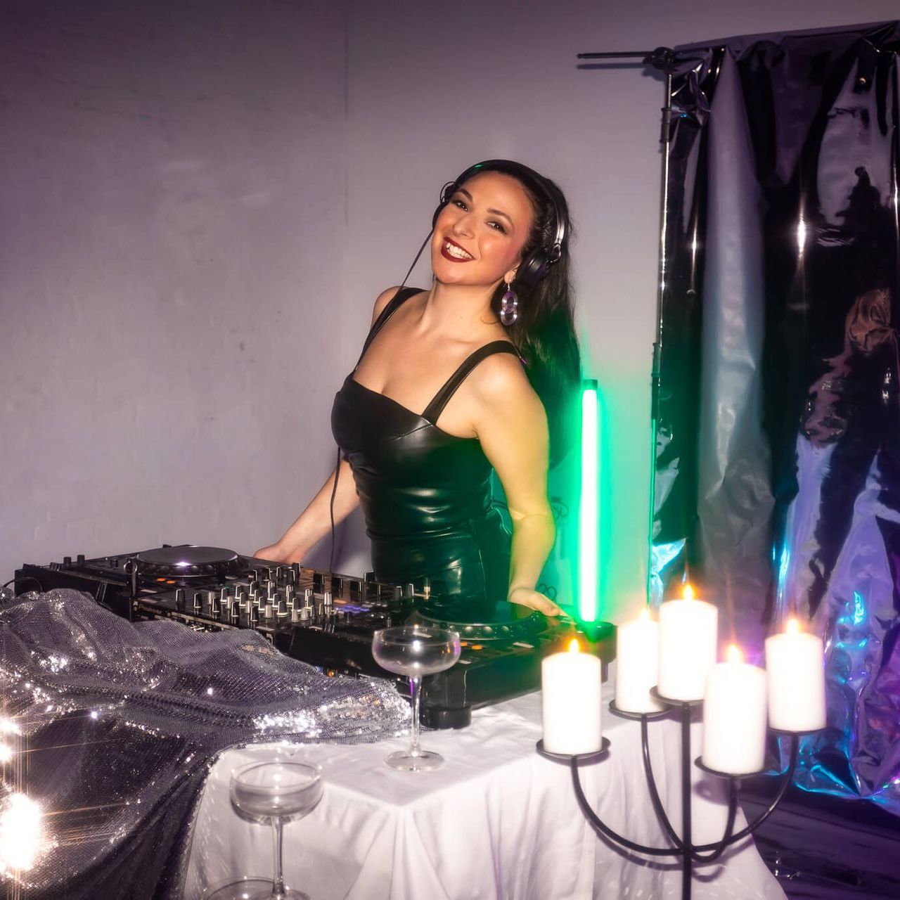 A smiling DJ wearing a dark outfit and headphones stands at a mixing console behind a table with candles.