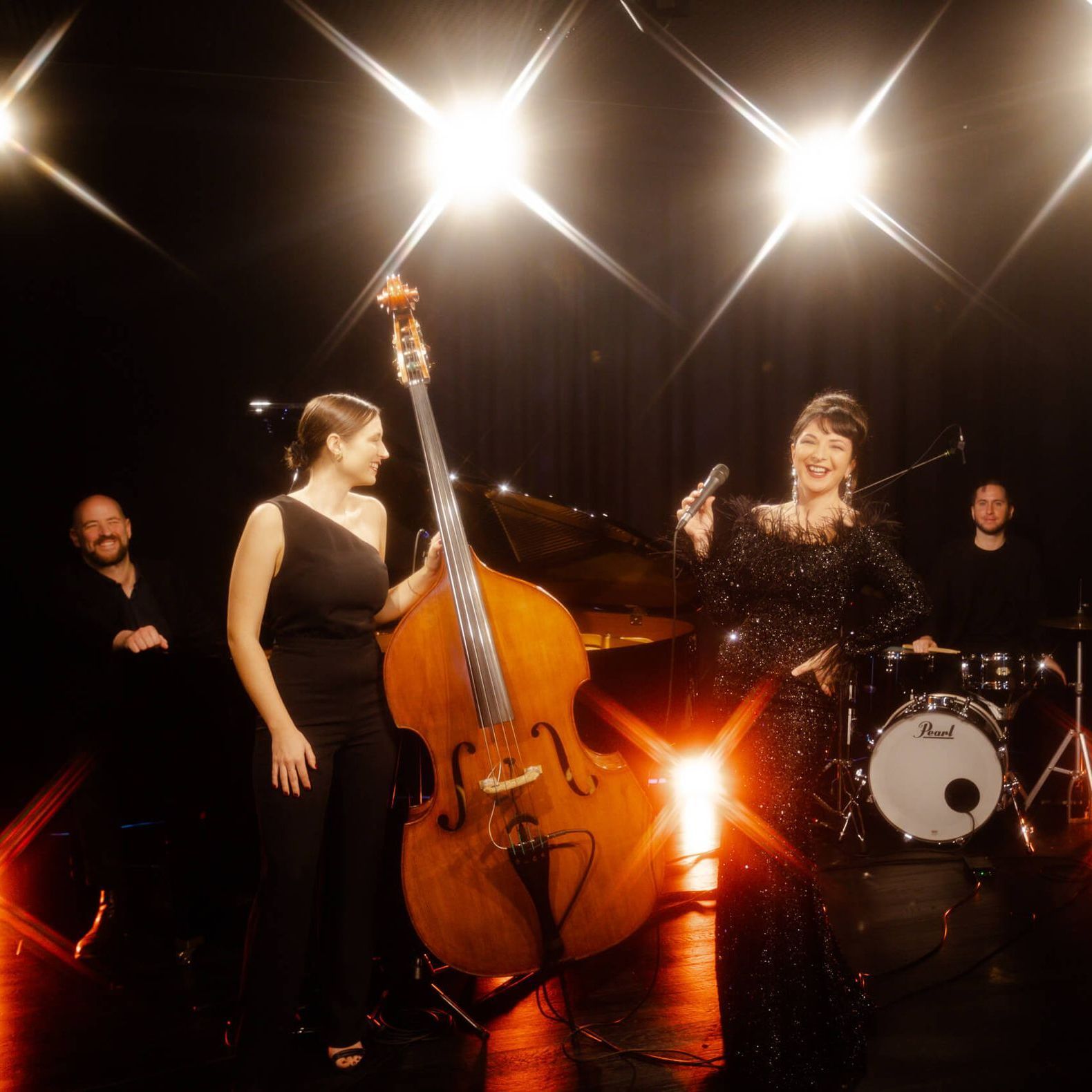 A musical quartet performs on a dimly lit stage with bright spotlights, featuring a bassist, singer, and drummer.