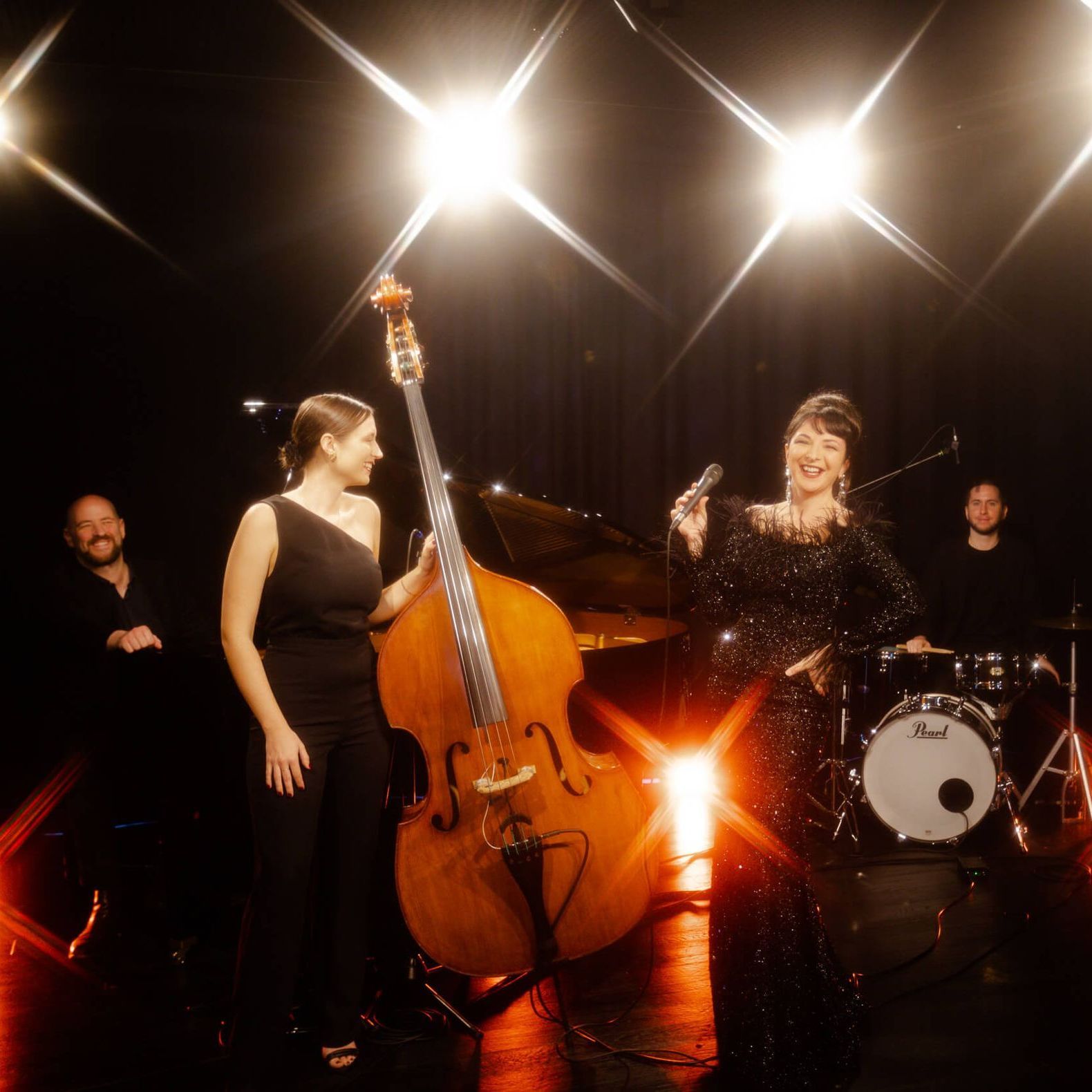 A jazz quartet performs on a stage, with a vocalist, upright bassist, pianist, and drummer under bright stage lights.
