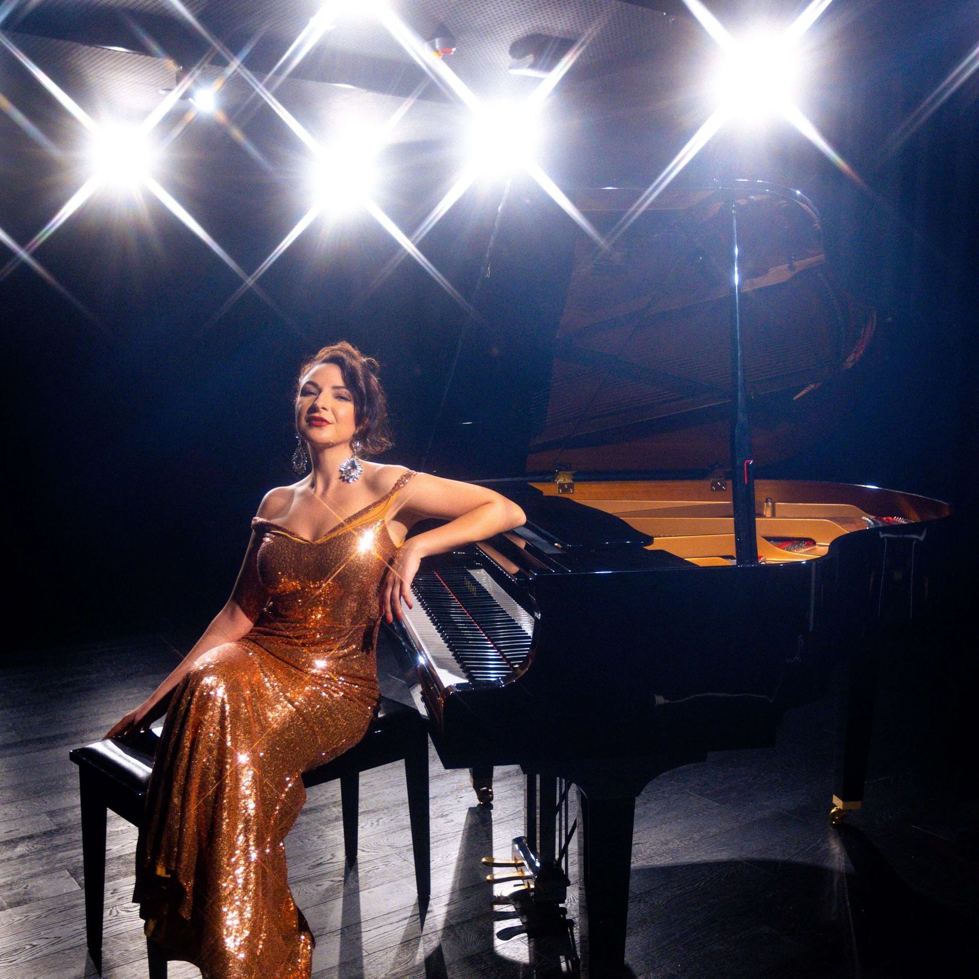 A person in a shimmering gold gown sits at a grand piano on a stage lit by bright, star-effect spotlights.