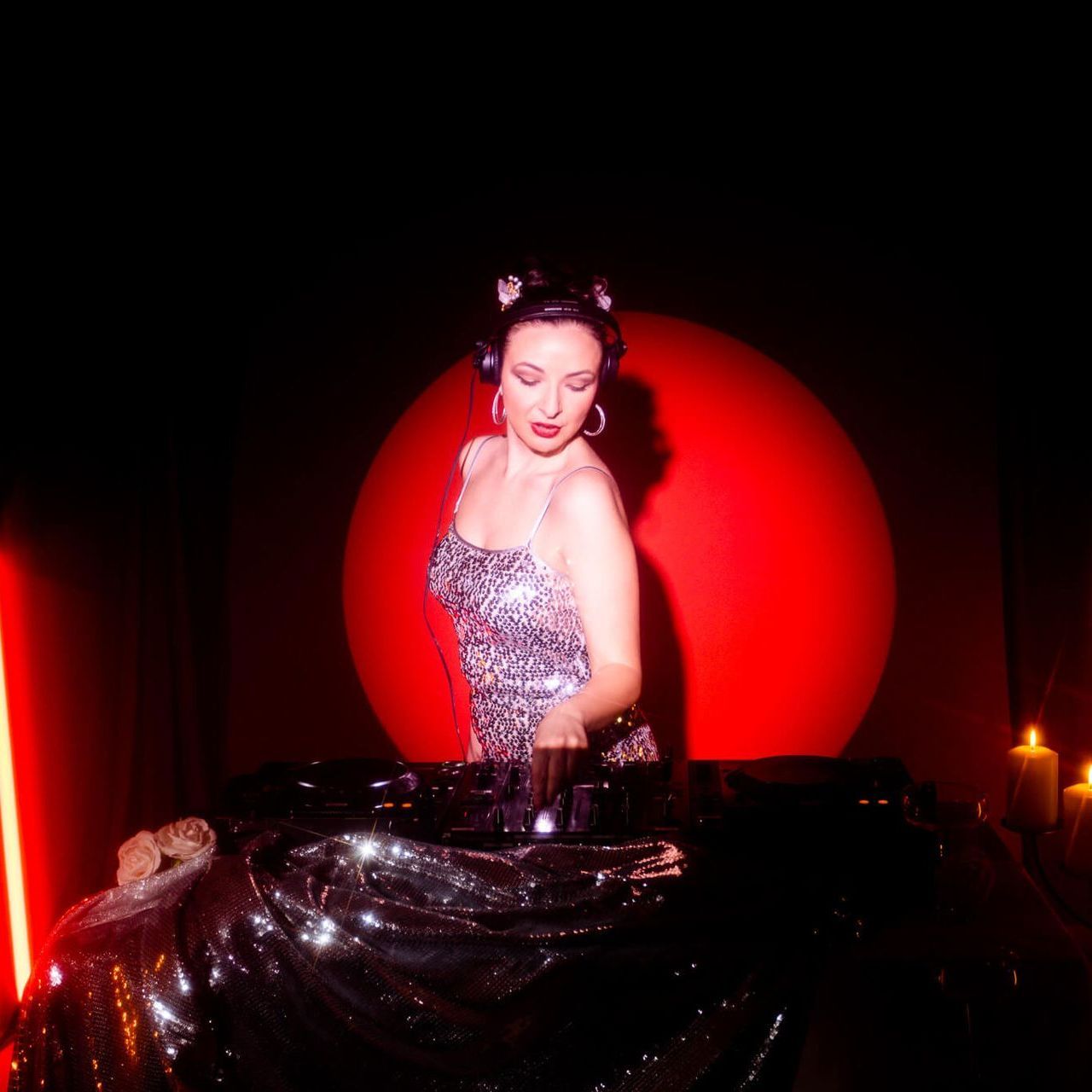 A DJ wearing a sequined top stands behind a dark, shimmering console against a backdrop of a large red circle.