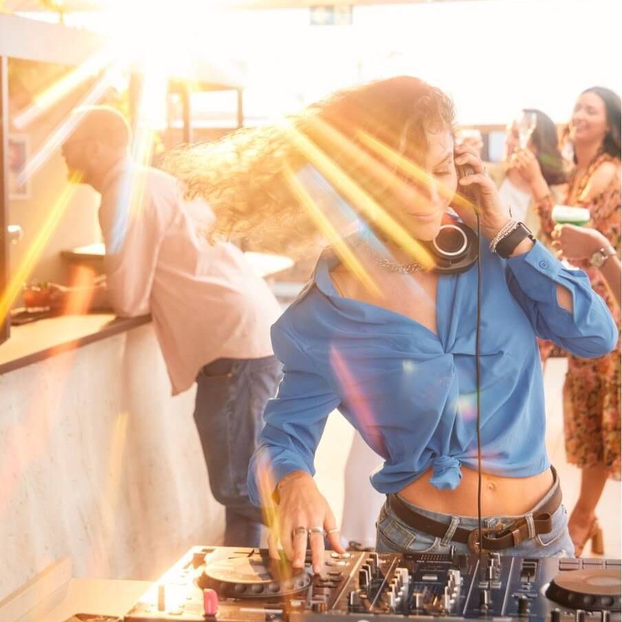 DJ wearing headphones at a party, mixing music. Sunlight beams. People in background.