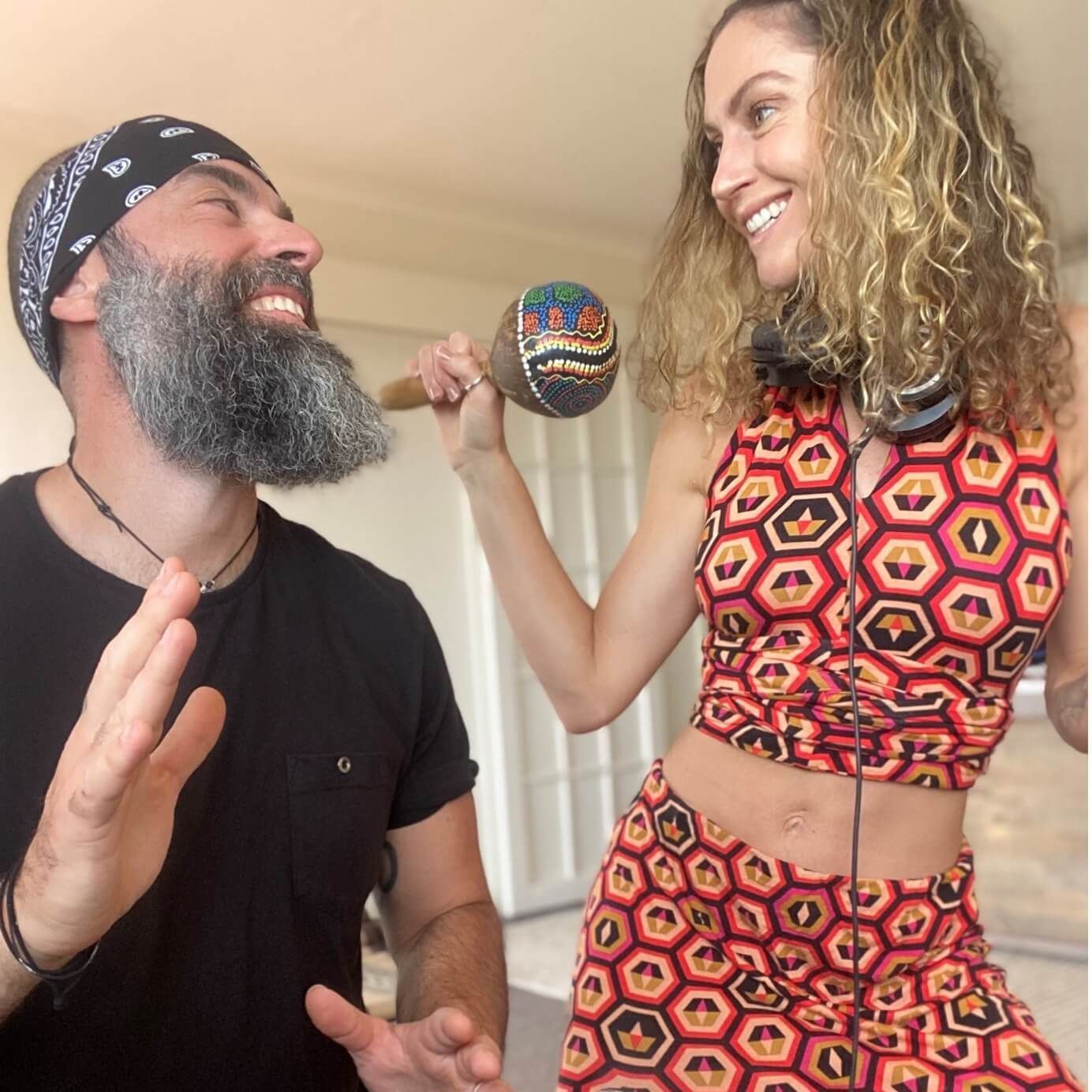 Man in bandana and woman in matching outfit laughing with a maraca indoors.