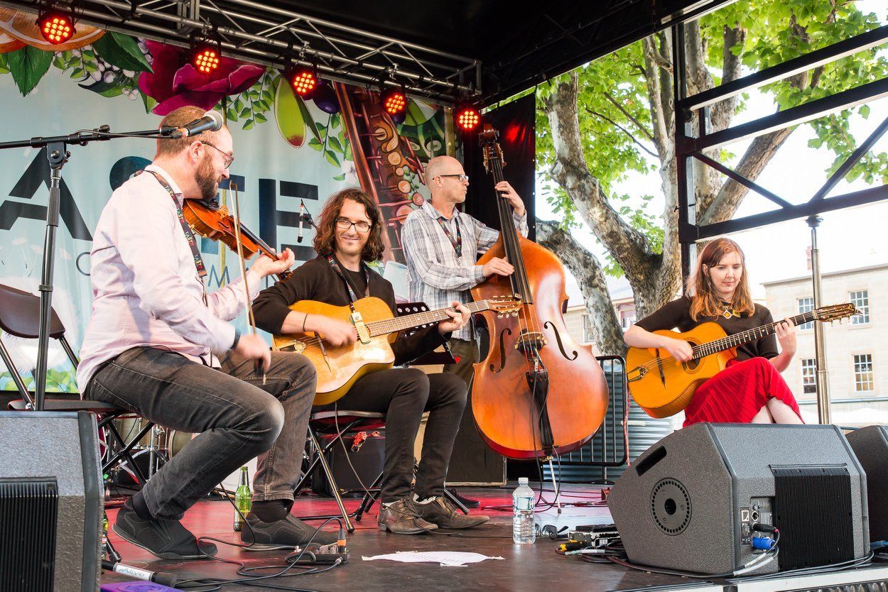 A string band performs on a stage outdoors. Musicians play violin, guitars, and upright bass under a banner.