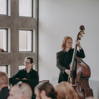 Two musicians perform, one playing a double bass, the other seated with glasses at a keyboard.
