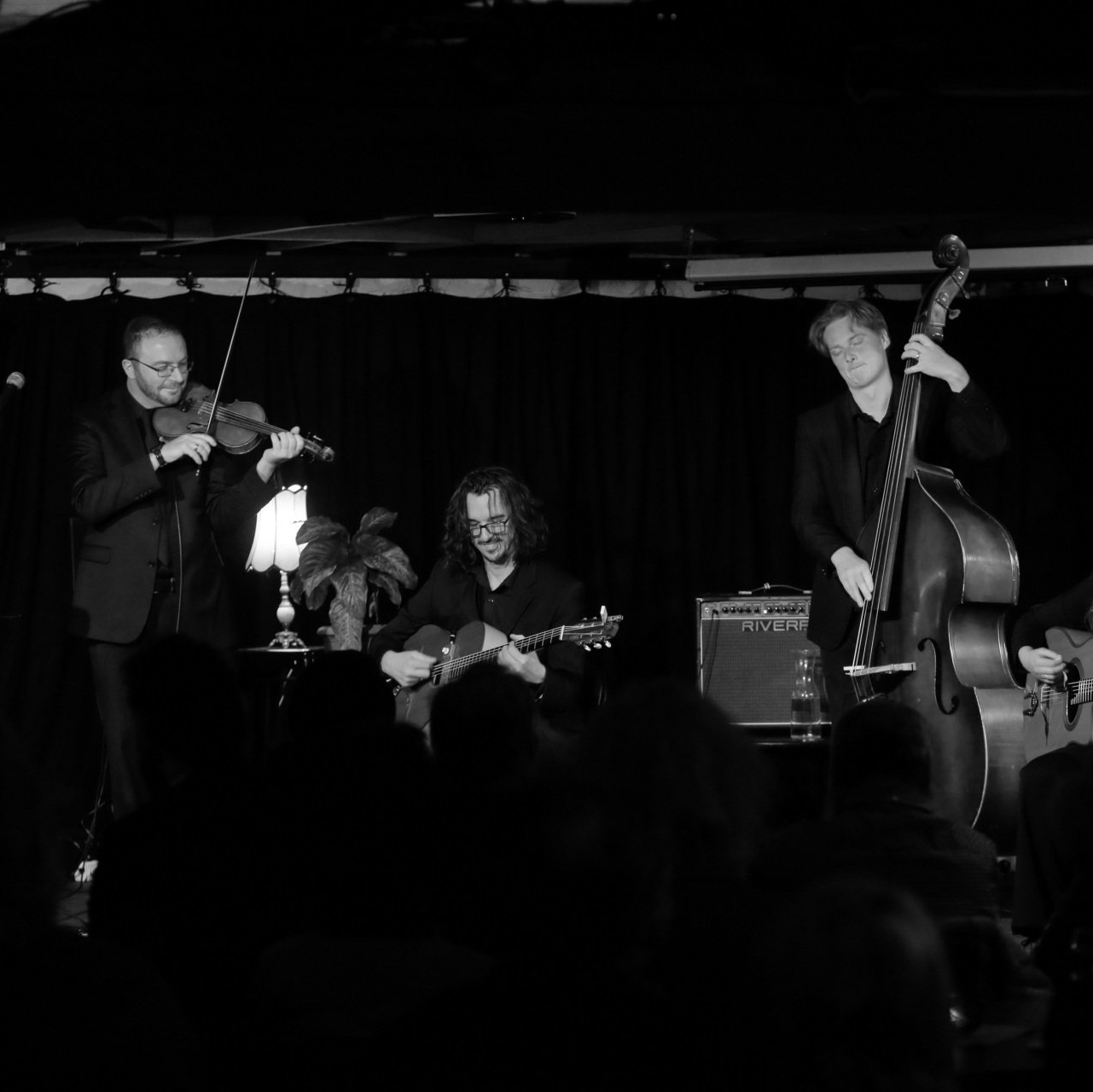 Band performing on stage: violinist, guitarist, bassist; black and white photo.