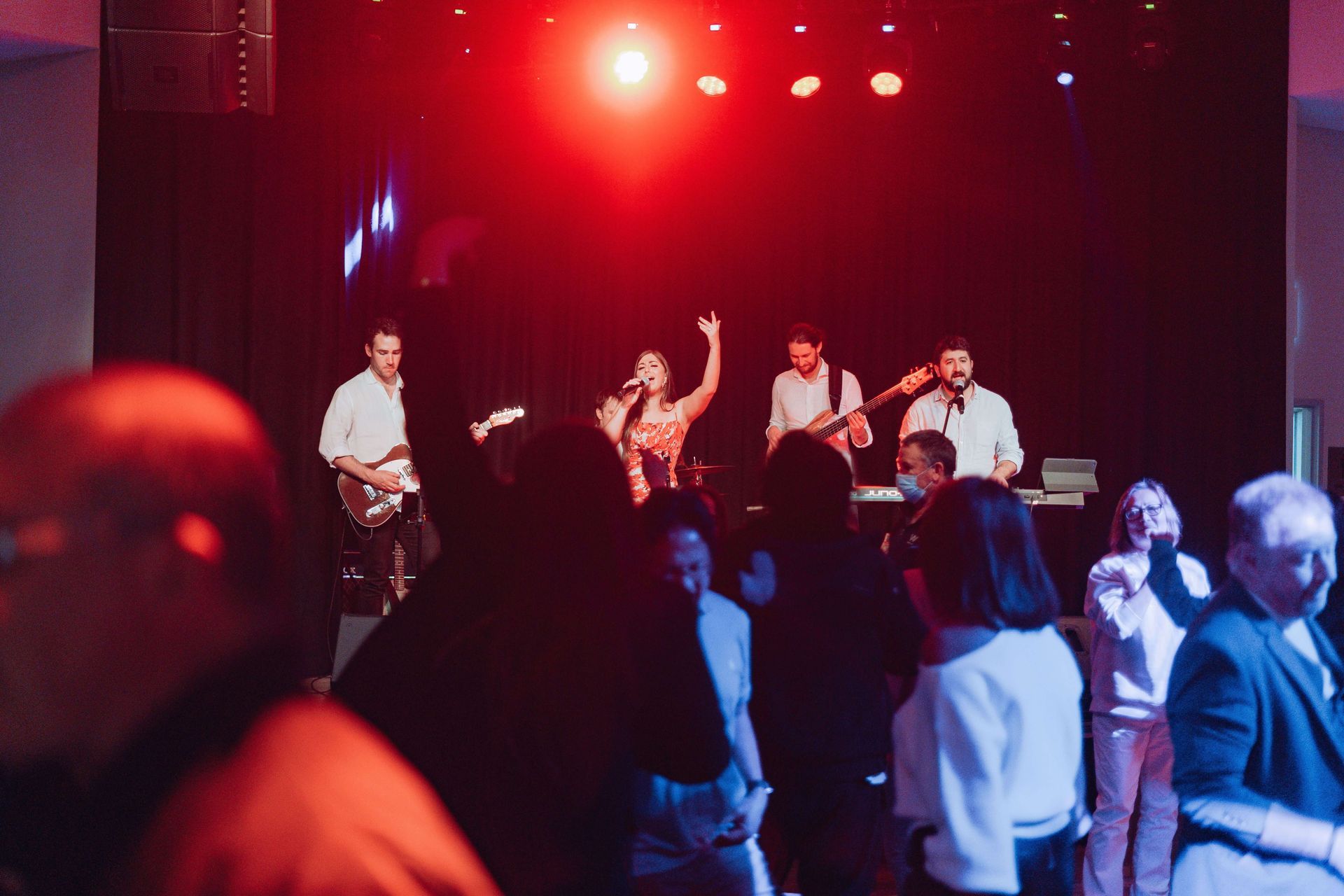 A band performs on a dimly lit stage with red lighting, while an audience dances in the foreground.