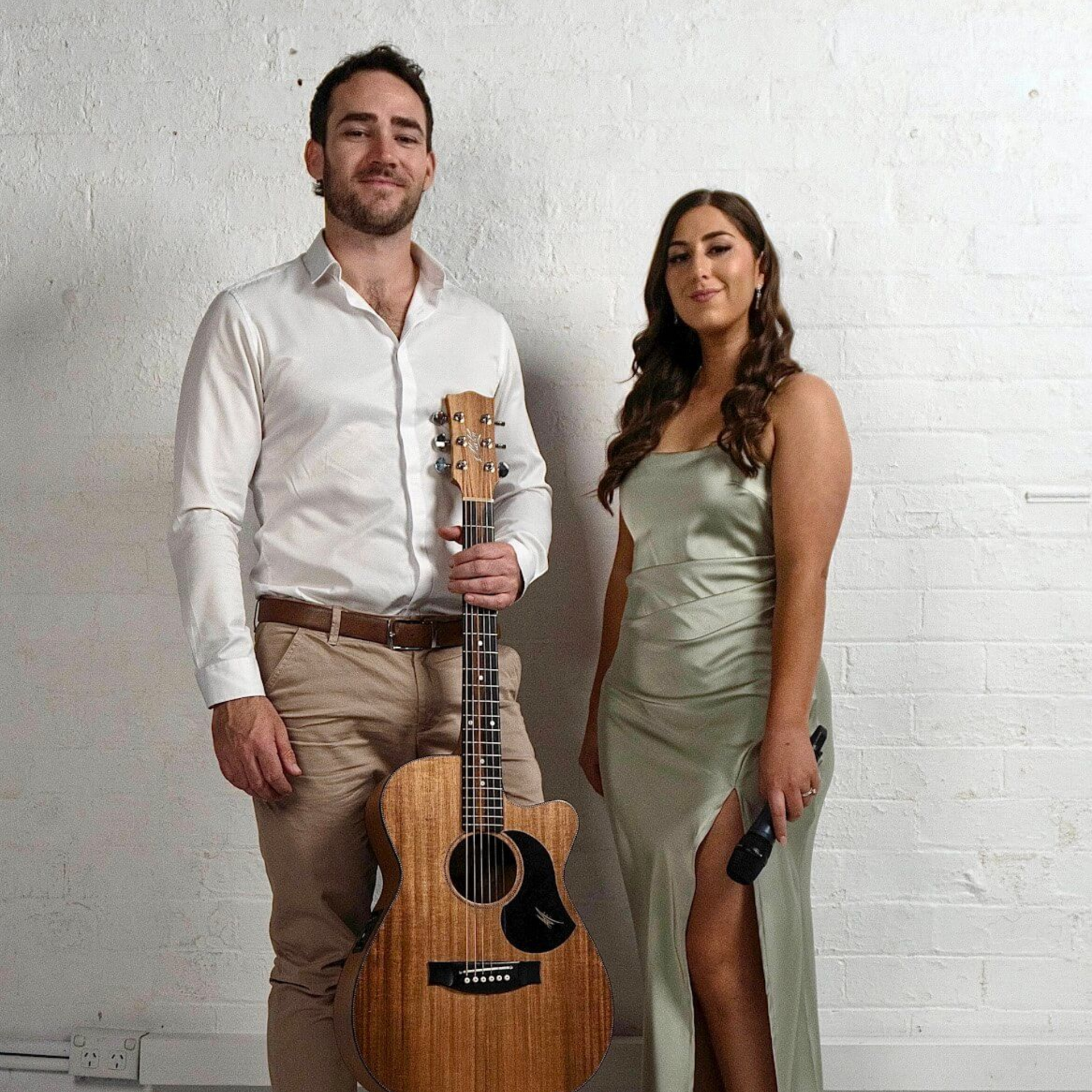 A man in a white shirt holding an acoustic guitar stands next to a woman in a sage green dress holding a microphone.