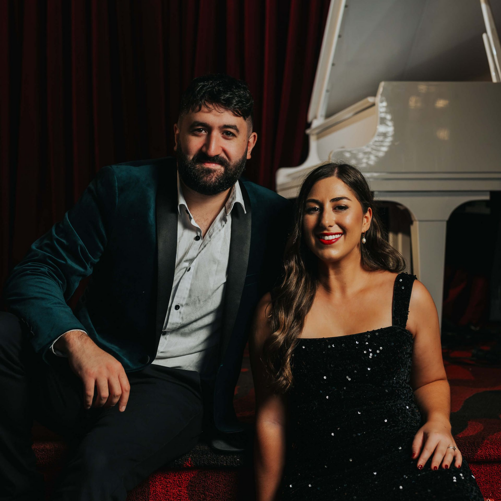 A man in a teal velvet blazer and a woman in a black sequined dress sit together in front of a white grand piano.