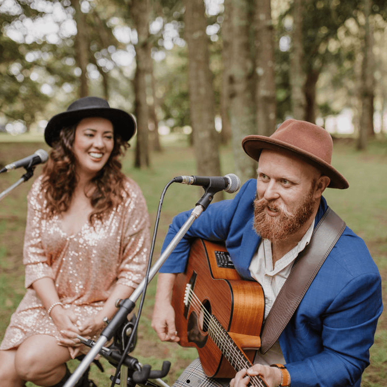 A musician with a red beard playing guitar and a woman in a sequined dress singing, both wearing hats in a forest setting.