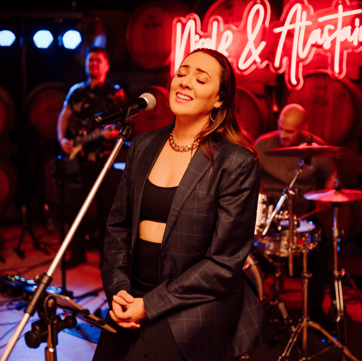 A singer performs into a microphone on stage, accompanied by a guitarist and a drummer in a dimly lit, red-hued venue.