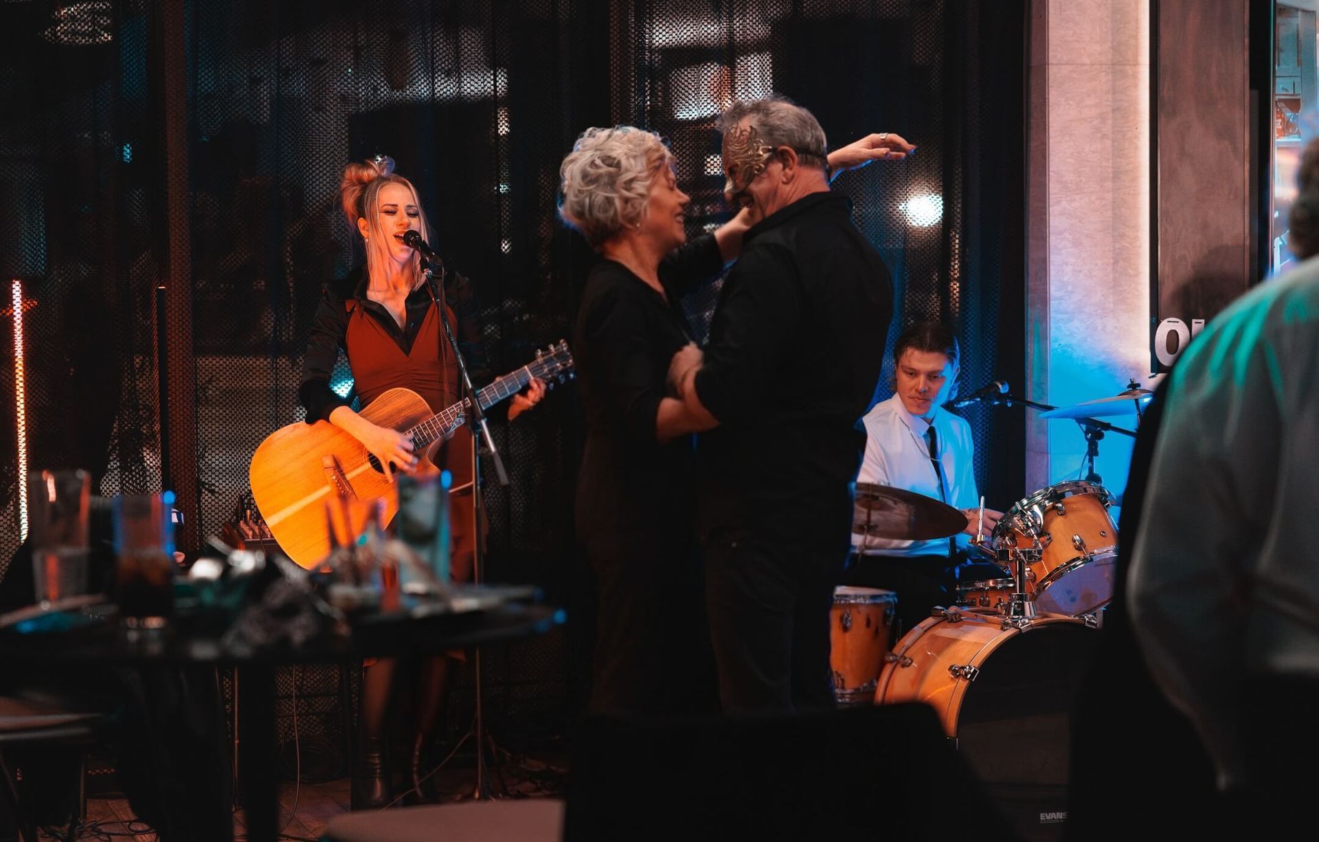 Live band performing on a dimly lit stage, with guitar, drums, and singers under warm orange and blue lights