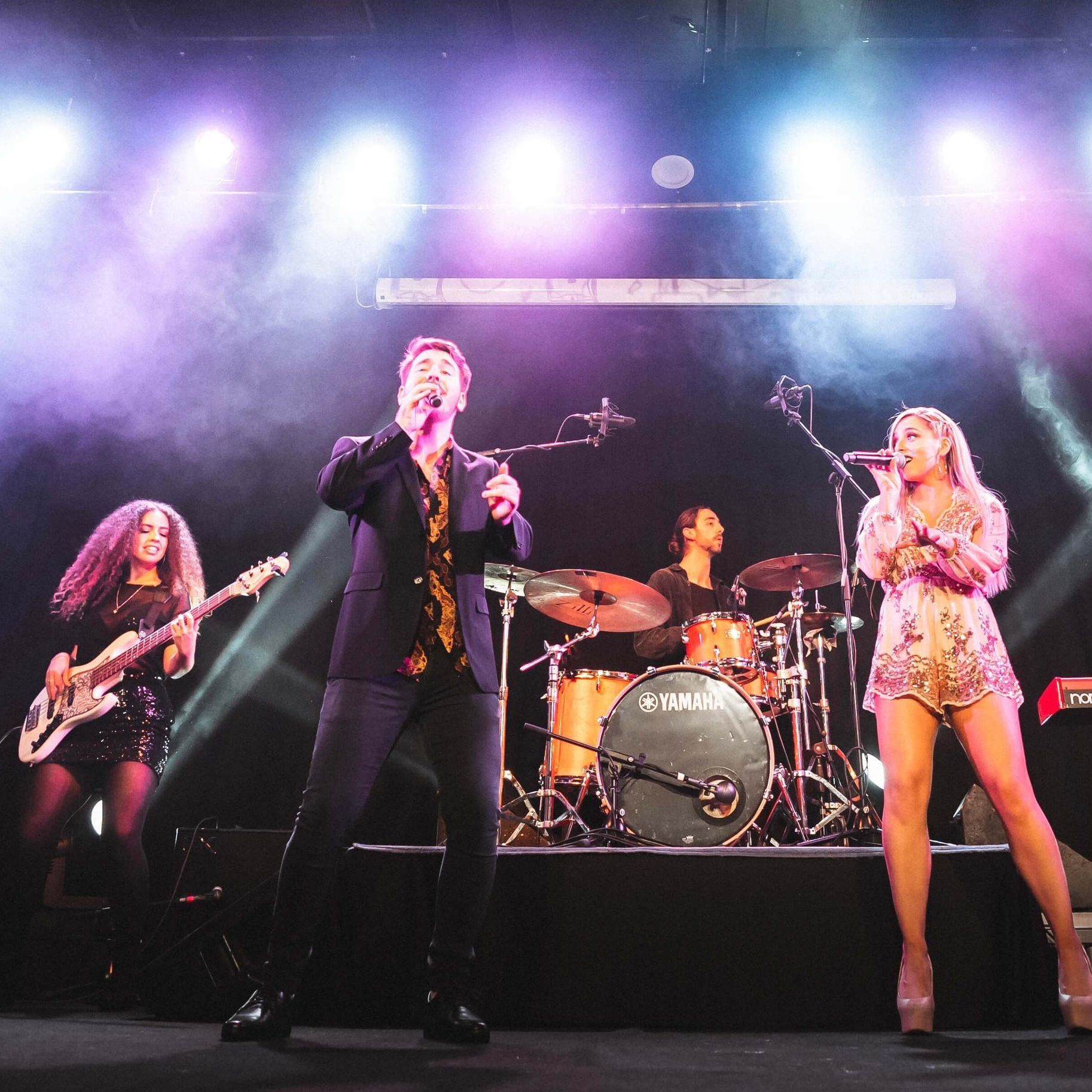 A band performing on a stage under bright spotlights, featuring two vocalists, a guitarist, and a drummer.