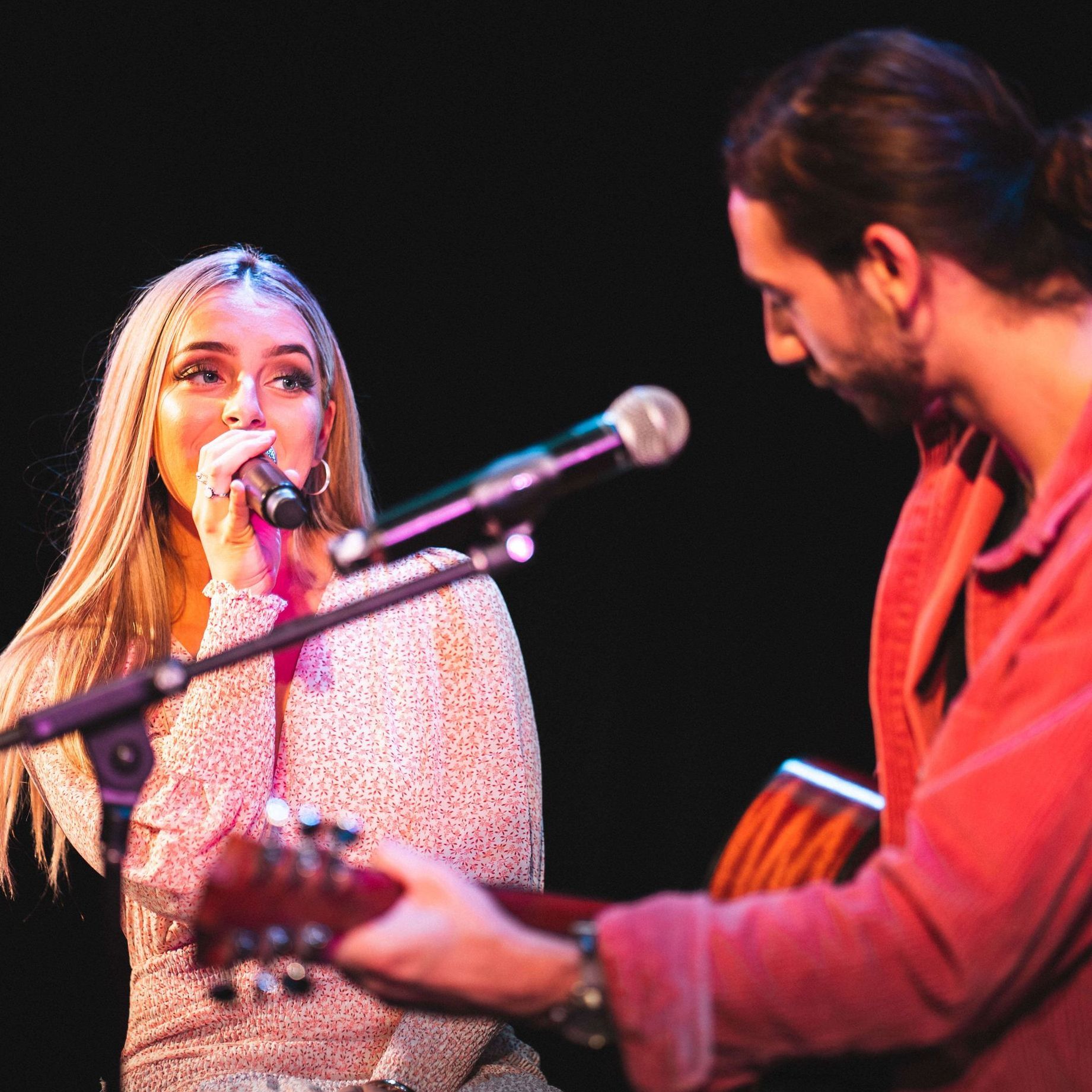A vocalist sings into a microphone while accompanied by an acoustic guitarist on a dimly lit stage.